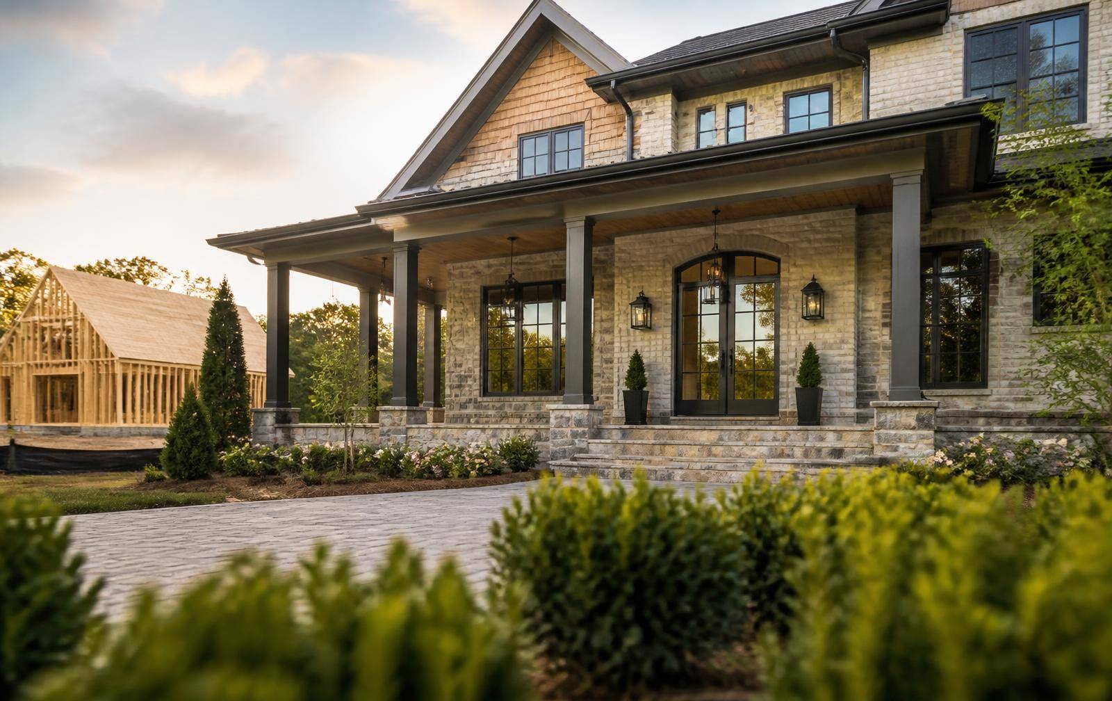Newly completed brick and stone luxury home at golden hour with a framed house under construction beyond